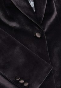 Blazer de terciopelo negro con una textura suave, solapa con muesca y tres botones negros en el puño. Presenta un forro interior con un diseño estampado.
