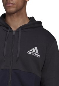 Sweat à capuche gris zippé avec un panneau plus foncé, des cordons ajustables et un logo Adidas blanc brodé sur la poitrine.