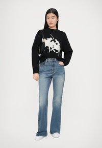 Črn pulover z grafiko belega igralca poloja, kombiniran s svetlo modrimi razširjenimi jeans hlačami. Model drži majhno črno torbico in nosi bele superge.