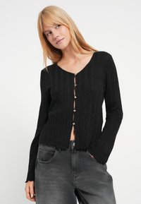 Μαύρο πλεκτό ζακέτα με λαιμόκοψη σε σχήμα V, μακριά μανίκια και έξι κουμπιά στο μπροστινό μέρος, με υφασμάτινο σχέδιο και κοντό τελείωμα.