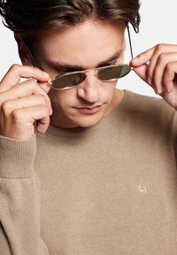 Okulary przeciwsłoneczne w złotej oprawie z ciemnozielonymi soczewkami trzymane przez osobę w beżowym swetrze, który ma subtelną wyszywaną metkę.