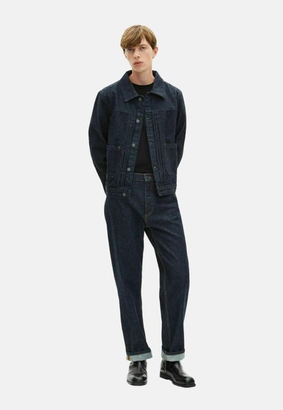 Chaqueta de mezclilla oscura con dos bolsillos en la parte frontal, combinada con pantalones de mezclilla oscuros a juego. Los puños están doblados y unas botas de cuero negro completan el look.