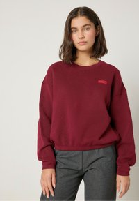 Burgundi spalvos džemperis su tekstūru, laisvas kirpimas, raukiniai rankogaliai ir mažas raudonas logotipas krūtinėje. Derinama su pilkais keliais.