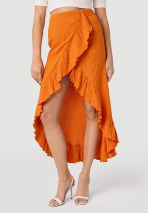Oranje wikkelrok met een gestructureerde afwerking, voorzien van een gerimpelde zoom en hoge split. Gepaard met witte hoge hakken sandalen.