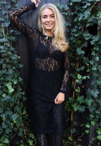 Fekete csipkeruha hosszú ujjakkal, virágmintákkal és áttetsző kialakítással. Nem átlátszó anyagra húzva, kiegészítve kültéri háttérrel.