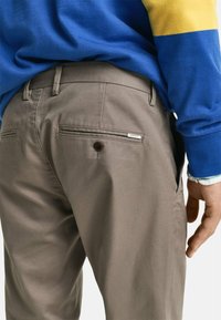Pantalones grises con una textura suave, que cuentan con dos bolsillos traseros, uno de ellos adornado con un botón marrón y un pequeño detalle de logo.