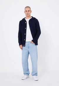 Marineblauer Strickcardigan mit Kapuze, verziert mit einem Rautenmuster und großen Knöpfen, kombiniert mit einem weißen T-Shirt und hellblauen Jeans.