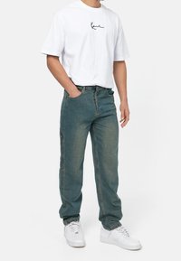 Vit T-shirt med kort ärm och svart text på bröstet, kombinerad med ljus teal baggy jeans och vita sneakers med elastiska ärmslut.