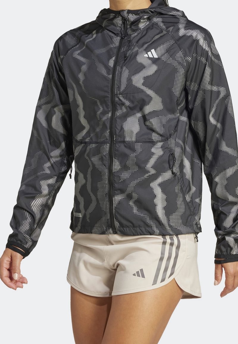 Femme portant une veste à capuche noire à motif avec fermeture éclair intégrale et un short de sport beige avec trois bandes sur le côté.