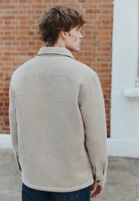 Beige textured shirt jacket with a collared neckline, buttoned cuffs, and a relaxed fit, paired with dark blue jeans.