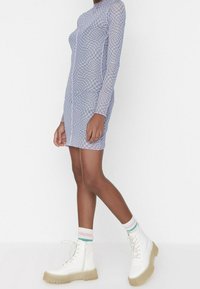 Robe cintrée à manches longues avec un motif à carreaux bleu clair et blanc, associée à des bottes blanches à lacets et des chaussettes rayées multicolores.