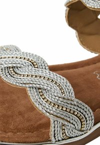 Des sangles en cuir tressées en argent avec des accents dorés sur une base en suède marron doux. Présente un design arrondi avec une semelle basse.