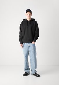 Hanorac negru cu buzunar kangaroo, asortat cu blugi ușor largi de culoare albastră deschisă și pantofi negri. Modelul poartă o șapcă închisă la culoare. Fundal simplu.
