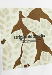 Medvilninis marškinėliai su dideliu mėtos žalumos lopu, kuriame yra rudos ir šviesiai smėlinės spalvos lapų raštas. Juodu tekstu parašyta "Originals Studio."