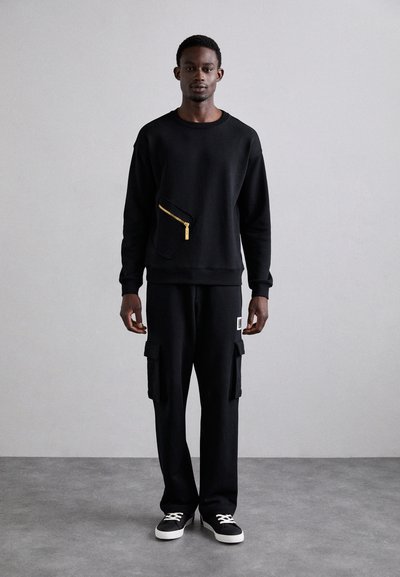 Čierna mikina so zlatým zipsovým vreckom; v kombinácii s čiernymi cargo nohavicami s bočnými vreckami. Model stojí na šedej podlahe.