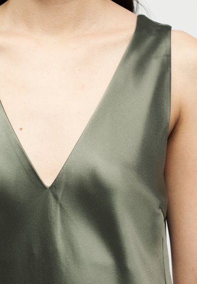 Κοντινό πλάνο ενός ατόμου που φοράει αμάνικο τοπ από σατέν ελιάς με βαθύ V-λαιμόκοψη, που αποκαλύπτει το πάνω μέρος του στήθους και τους ώμους.