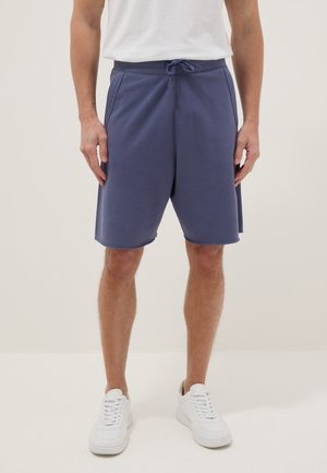 Hombre vestido con pantalones cortos azules hasta la rodilla con cordón, zapatillas blancas y una camisa blanca, de pie frente a un fondo claro y simple.