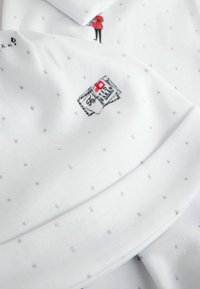 Vit bomullsklädsel med mönster av små bokstäver. Inkluderar en vikt krage och en broderad tygmärke med ett bagarmotiv.