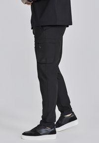 Pantalones cargo negros fabricados con tela resistente, que cuentan con un bolsillo lateral, puños elásticos y un pequeño detalle de logo. Combinados con zapatillas negras.