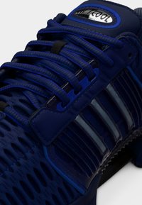 Μπλε αθλητικό παπούτσι με υφασμάτινο επάνω μέρος με υφή, πολυστρωματικές επιφάνειες, χοντρά κορδόνια και γυαλιστερή λεπτομέρεια. Εμφανές λογότυπο στη γλώσσα του παπουτσιού.