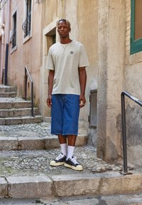 Beige t-shirt med en solgrafik, blå denimshorts og sorte sneakers med hvide detaljer, sat mod en brostensbelagt gade.