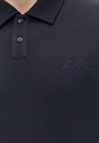 Gros plan sur un polo bleu marine foncé avec deux boutons noirs et "Les Deux" brodé avec un fil assorti sur la poitrine.