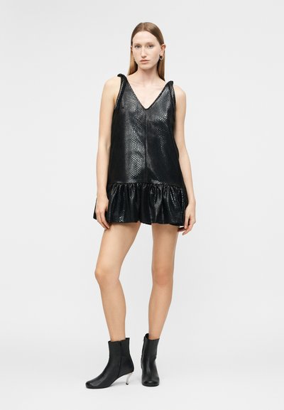 Młoda kobieta ubrana w czarną, bezrękawną, teksturowaną mini sukienkę z falbaną u dołu i czarne botki, stojąca na tle jednolitego szarego tła.