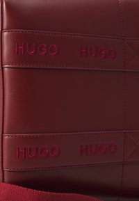Raudona odinė rankinė su iškilniu "HUGO" prekės ženklu ir kontrastingomis siūlėmis, turinti tekstūruotą paviršių ir stačiakampį dizainą.