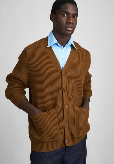 Brun ribbet cardigan med V-hals, frontknapper og to store lommer, båret over en lyseblå skjorte med krave.