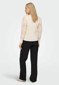 Cremefarvet top med gennemsigtige, voluminøse blondeærmer, parret med sorte, brede bukser. Toppen har en rund halsudskæring og en tætsiddende design.