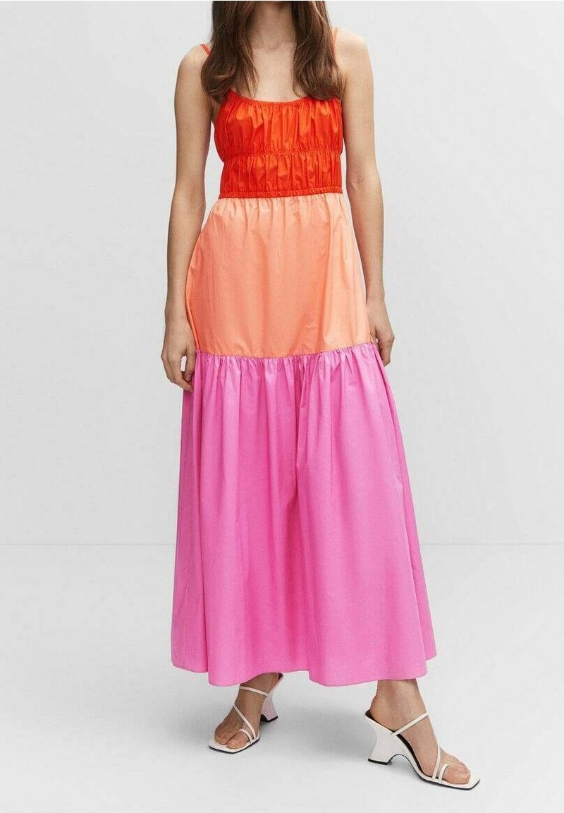 Femme portant une robe longue sans manches à volants avec des sections rouges, orange et roses, associée à des sandales blanches à talons hauts avec brides.