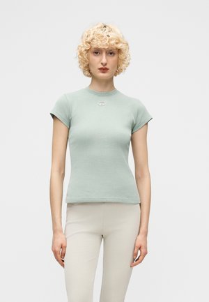 Donna con capelli biondi ricci che indossa una maglietta aderente a maniche corte verde chiaro e pantaloni beige chiaro, in piedi davanti a uno sfondo neutro.