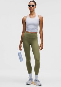El atuendo atlético presenta una camiseta sin mangas gris claro con aberturas en los hombros y leggings de talle alto en verde olivo. Los accesorios incluyen gafas de sol y una botella de agua.