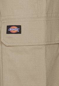 Béžová pracovní tkanina s mřížkovou texturou, s výraznou černou nášivkou loga Dickies na kapse. Šití viditelné po okrajích.