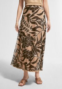 Jupe longue à motif floral dans des tons beige et marron, avec une coupe fluide et une ceinture à la taille. Associée à des sandales à talons marron.
