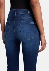 Jeans en denim bleu foncé au design ajusté, avec cinq poches, des coutures contrastantes et une étiquette de marque sur la taille et la poche.