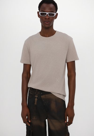 Beige t-shirt med rund hals i mjukt material, med en liten logotyp på vänster sida. Bärs med mörka mönstrade cargobyxor.