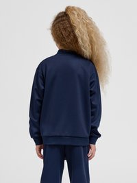 Enfant aux cheveux bouclés volumineux, moitié noirs, moitié blonds, de dos, portant un survêtement bleu marine sur un fond gris uni.