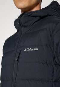 Muškarac u crnoj jakni od puffy materijala s kapuljačom, prednjim zatvaračem, zatvorenim džepom na prsima i bijelim Columbia logom na lijevoj strani prsnog koša.