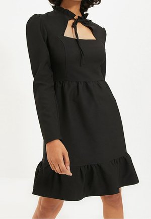 Robe noire à manches longues avec encolure carrée, col à volants noué par un ruban, taille froncée et ourlet à volants au-dessus des genoux.