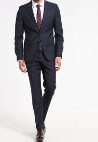 Marineblauw pak met een ingesneden revers, een knoopssluiting met twee knopen en een slim fit. Witte blouse eronder, bordeauxrode stropdas met stippen en bruine nette schoenen.