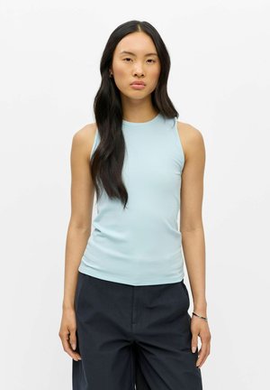 Jeune femme aux longs cheveux noirs portant un haut bleu clair sans manches et un pantalon foncé, debout devant un fond blanc uni.