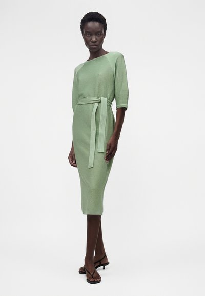 Γυναίκα που φοράει ένα ανοιχτοπράσινο midi φόρεμα με ριμπ ύφανση, μανίκια τρία τέταρτα και ταιριαστή ζώνη, συνδυασμένο με μαύρα πέδιλα με λουράκια.