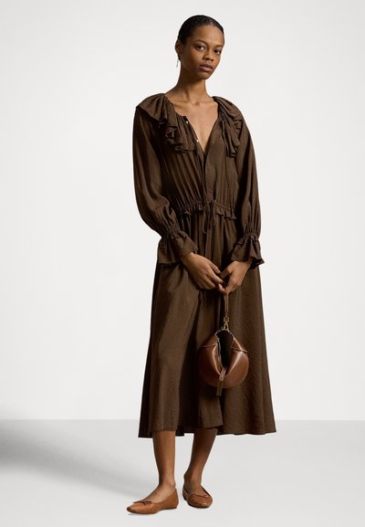 Ruda, ilgomis rankovėmis suknelė su raukinuku ant apykaklės ir elastine juosmens dalimi. Lygios tekstūros. Modelis laiko apvalią rudą rankinę ir dėvi lygias basutes.