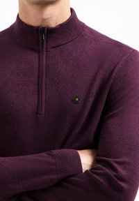 Bordeauxrotfarbener Pullover mit Reißverschluss aus weichem Stoff, mit geripptem Kragen und Bündchen sowie einem kleinen sechseckigen Logo auf der Brust.