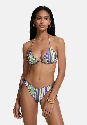 Donna con capelli lunghi e neri indossa un bikini a righe verdi, viola e bianche, orecchini a cerchio dorati e un braccialetto dorato spesso al polso destro.