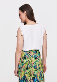 Biała bluzka z troczkami na ramionach i dekoracyjnymi koralikami, zestawiona z kolorową, wzorzystą zieloną spódnicą z tropikalnymi liśćmi.