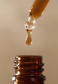 Bernsteinfarbener Flüssigkeitsstropfen, der von einem Glas-Tropfer in den offenen Flaschenhals einer braunen Flasche fällt, vor einem schlichten Hintergrund.