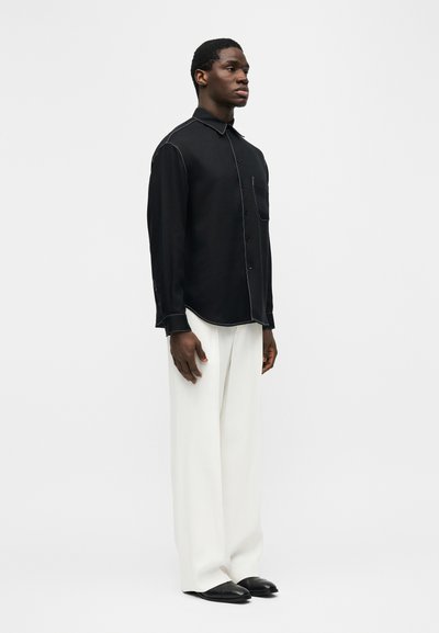 Άνδρας που στέκεται, φοράει μαύρο πουκάμισο με μακριά μανίκια και κουμπιά, με λευκά ραφτά, λευκό παντελόνι με φαρδιά πόδια και μαύρα παπούτσια, μπροστά σε απλό φόντο.
