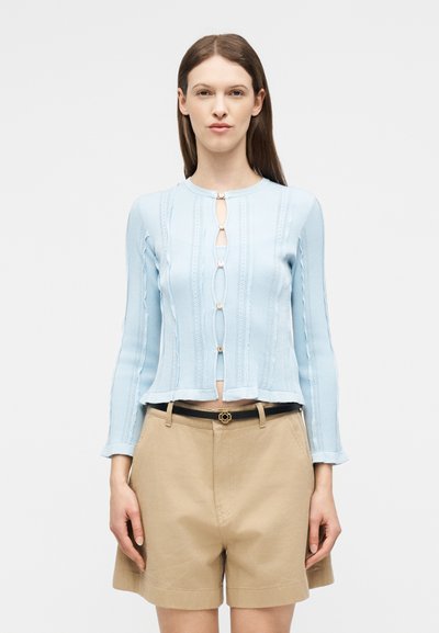 Γυναίκα που φορά ανοιχτό μπλε πλεκτό ζακέτα με διακοσμητικά κουμπιά και μπεζ σορτς ψηλόμεσα, στέκεται μπροστά σε απλό φόντο.